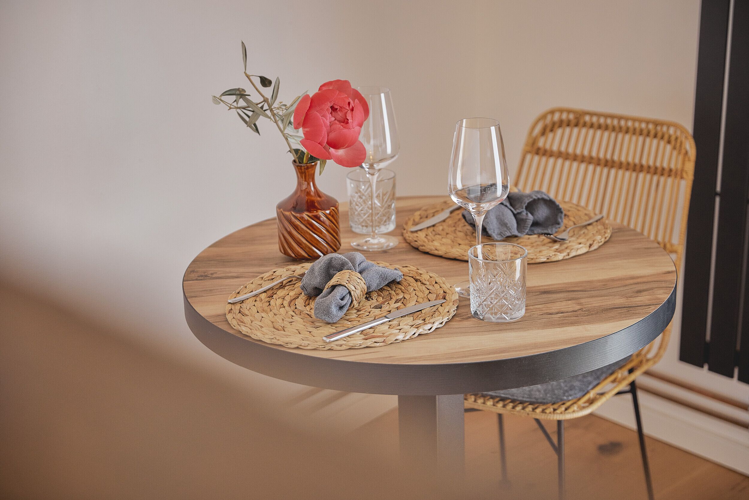 A round wooden table with woven placemats, a vase with a pink flower, and two wine glasses.