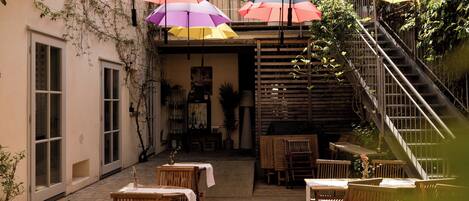 An outdoor patio area with wooden tables and chairs, covered by colorful umbrellas, and a staircase leading to an upper level.