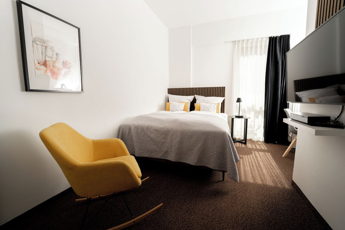 Een hotelkamer met een bed, een gele fauteuil, een bureau met een flatscreen televisie en een lijst aan de muur.