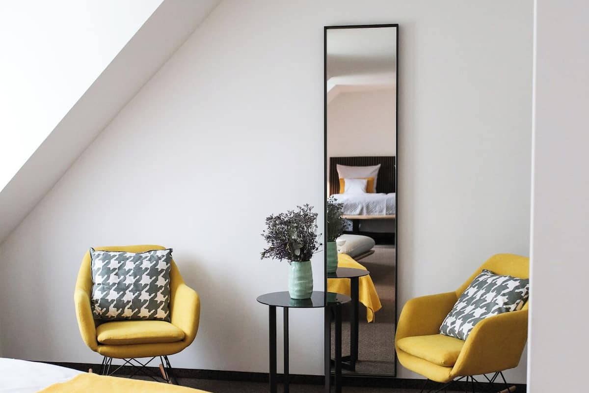 Een moderne hotelkamer met een schuine plafond, twee gele fauteuils, een groot bed met een gele deken, een klein rond tafeltje met een vaas, en een grote spiegel aan de muur.