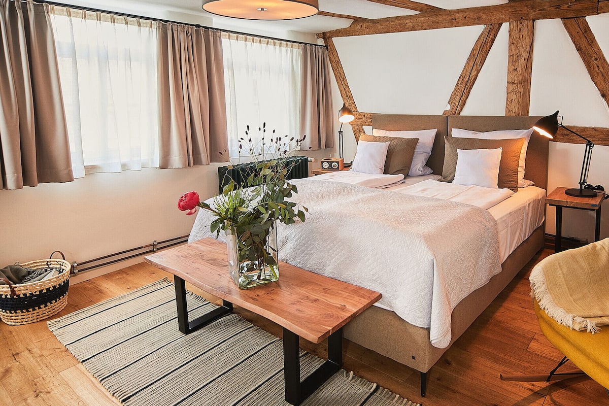 Een slaapkamer met een bed, nachtkastje, lamp, stoel en een raam met gordijnen.
