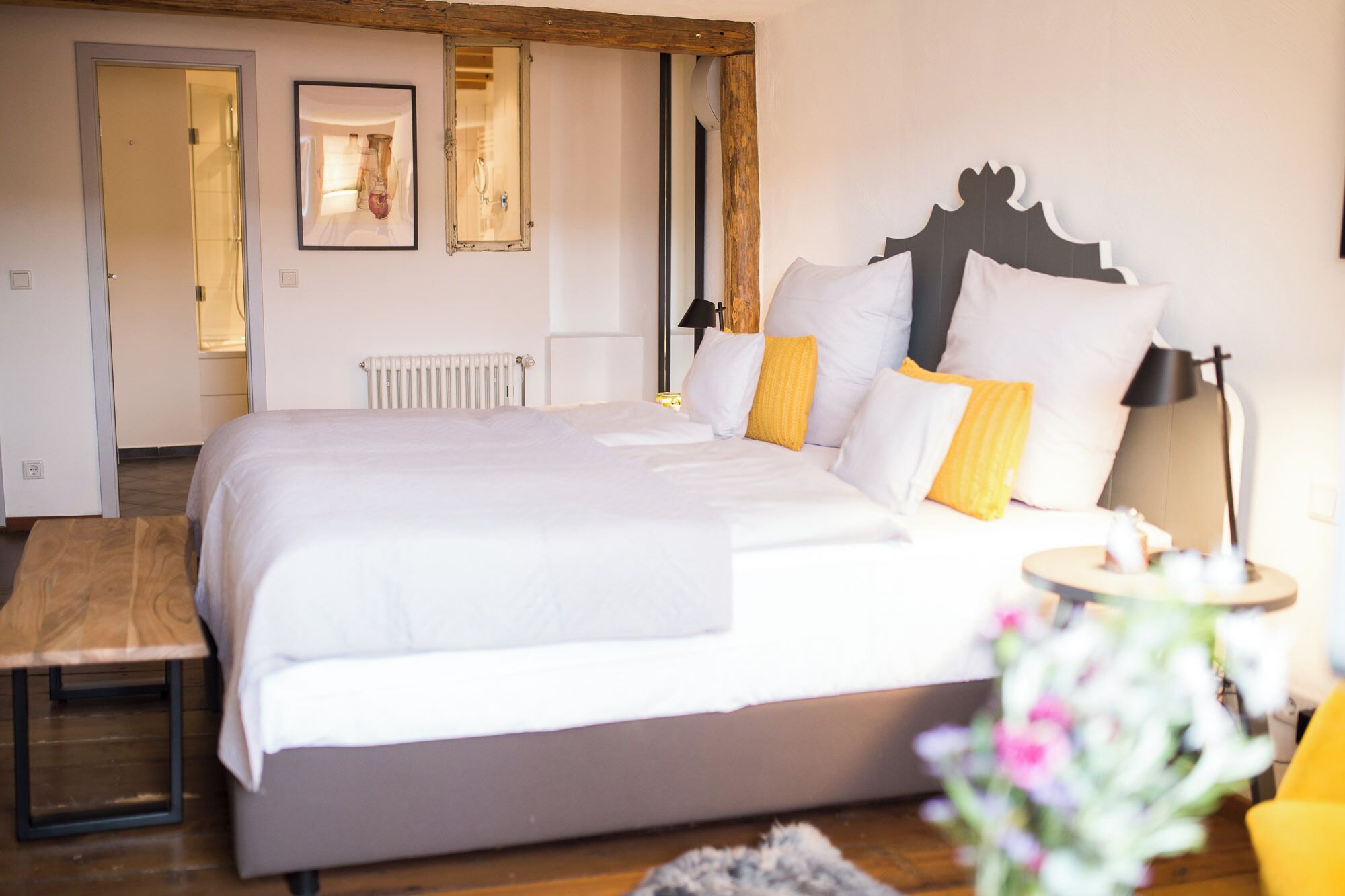 A bedroom with a large bed, a wooden bench, a bedside table, a mirror, and a radiator.