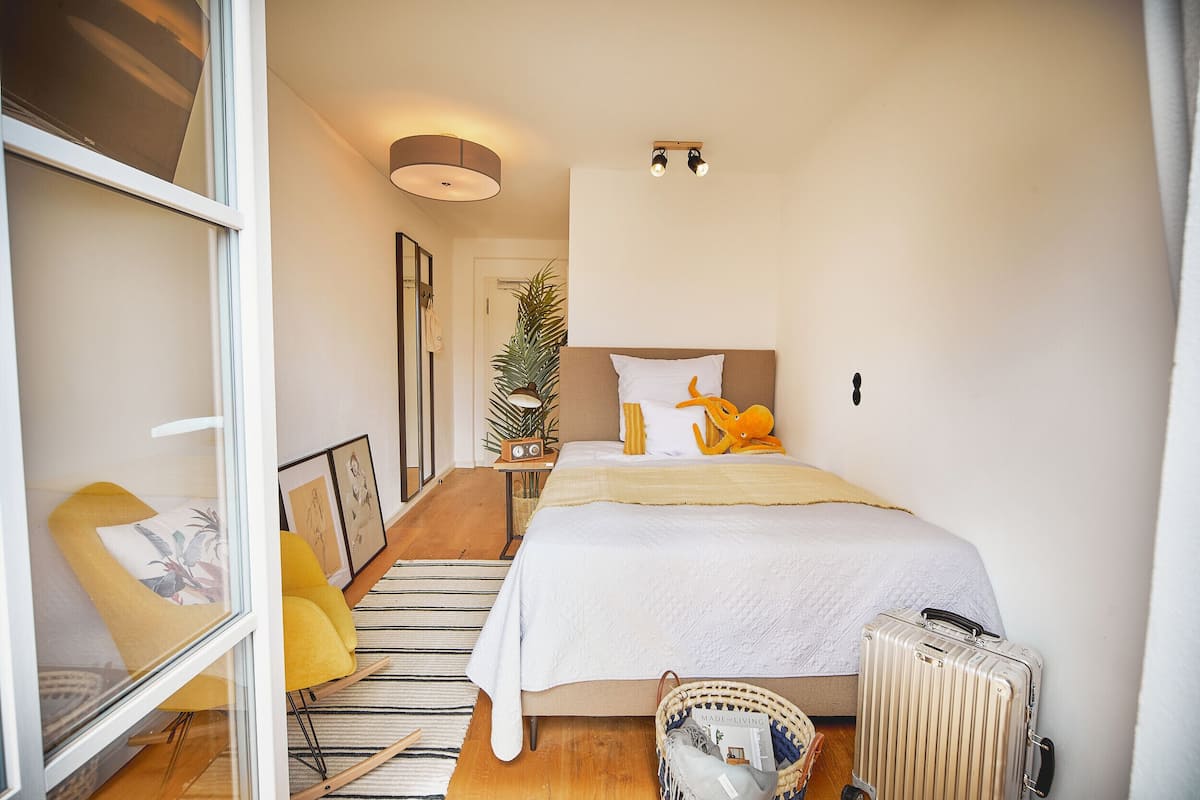 Een slaapkamer met een bed, een koffer, een radiator, een plant en een stoel.