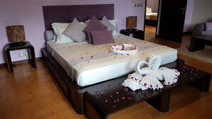 Uma cama bem arrumada com toalha e pétalas de rosa, uma mesa de cabeceira e vista para outro quarto.