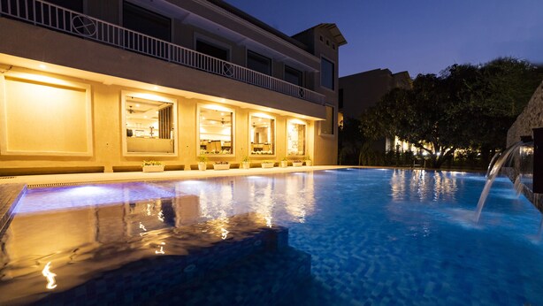 A swimming pool with a water feature, a building with multiple windows and balconies, and a night sky. - La Savanna (Ramnagar)