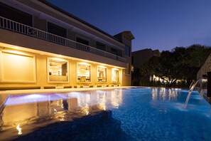 A swimming pool with a water feature, a building with multiple windows and balconies, and a night sky. - La Savanna (Ramnagar)