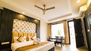 A hotel room with a large bed, a ceiling fan, a dining area with a table and chairs, and a view of the outdoors. - La Savanna (Ramnagar)
