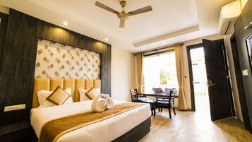 A hotel room with a large bed, a ceiling fan, a dining area with a table and chairs, and a view of the outdoors.