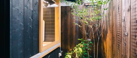 Un mur extérieur en bois, avec une fenêtre et un petit espace jardin agrémenté de plantes et d’un chemin en pierre.