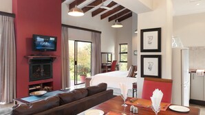 A hotel room with a large bed, a TV mounted on a red wall, a dining area with a wooden table set for two, and a living area with a brown sofa.
