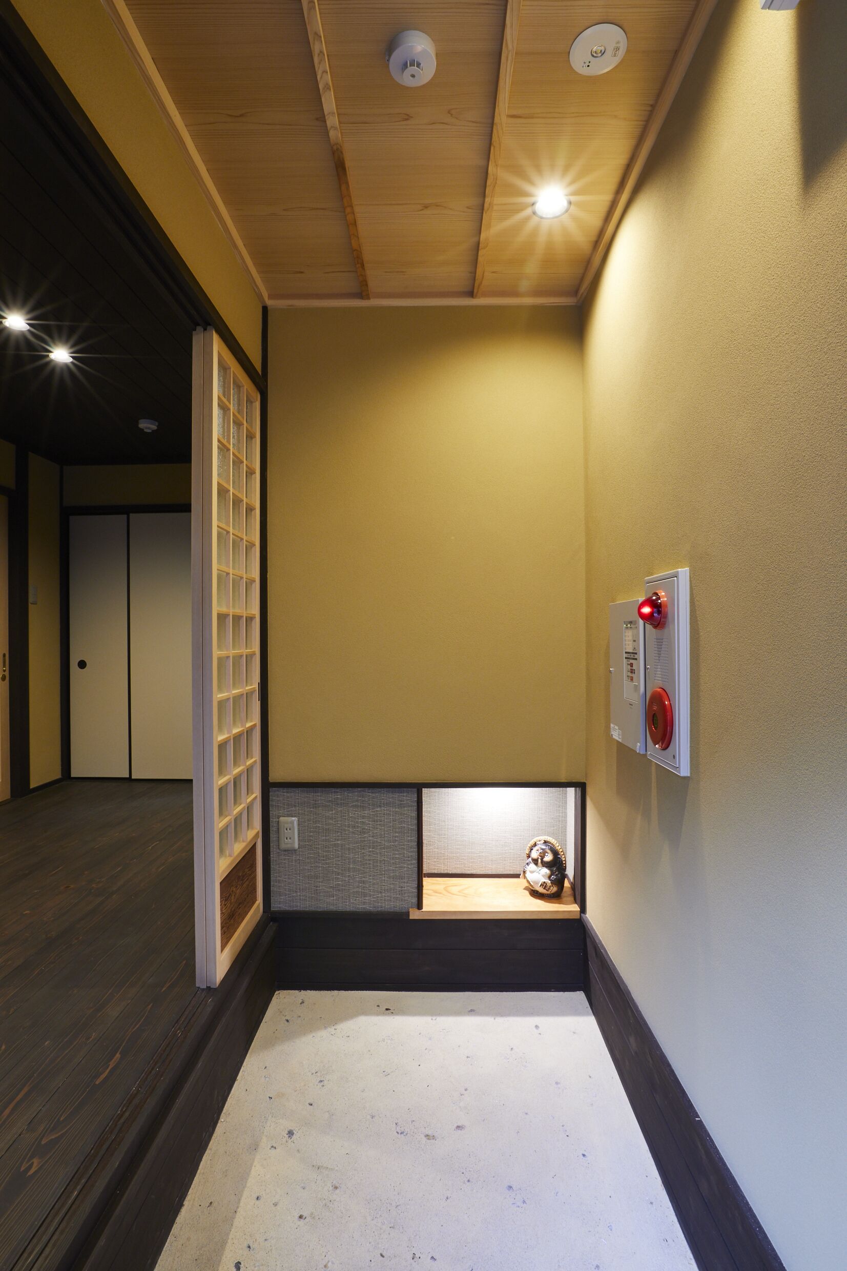 Um corredor com piso de madeira, teto rebaixado com iluminação, detector de incêndio na parede e uma pequena prateleira com um objeto decorativo.