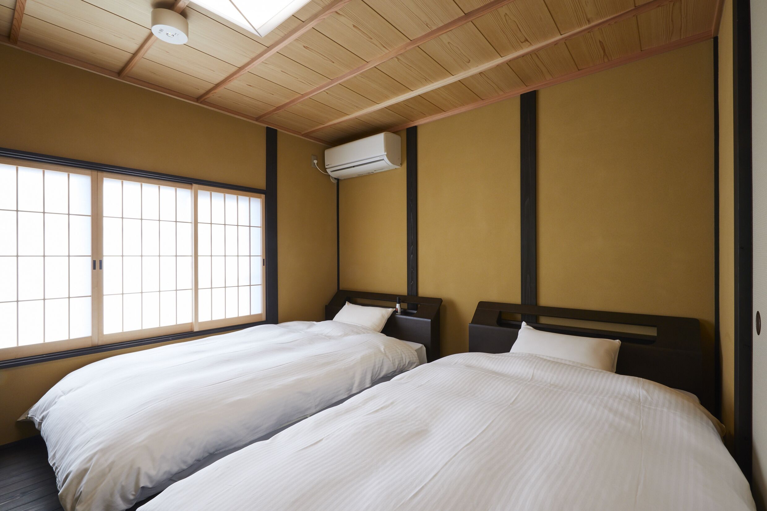 A hotel room with two beds, a ceiling fan, an air conditioning unit, and a large window with wooden frames.