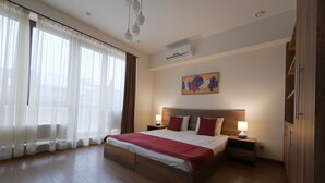 A hotel room with a large bed, wooden flooring, a nightstand with a lamp, a wall-mounted air conditioner, and a painting above the bed. - Welcome City Center Apartments (Yerevan)