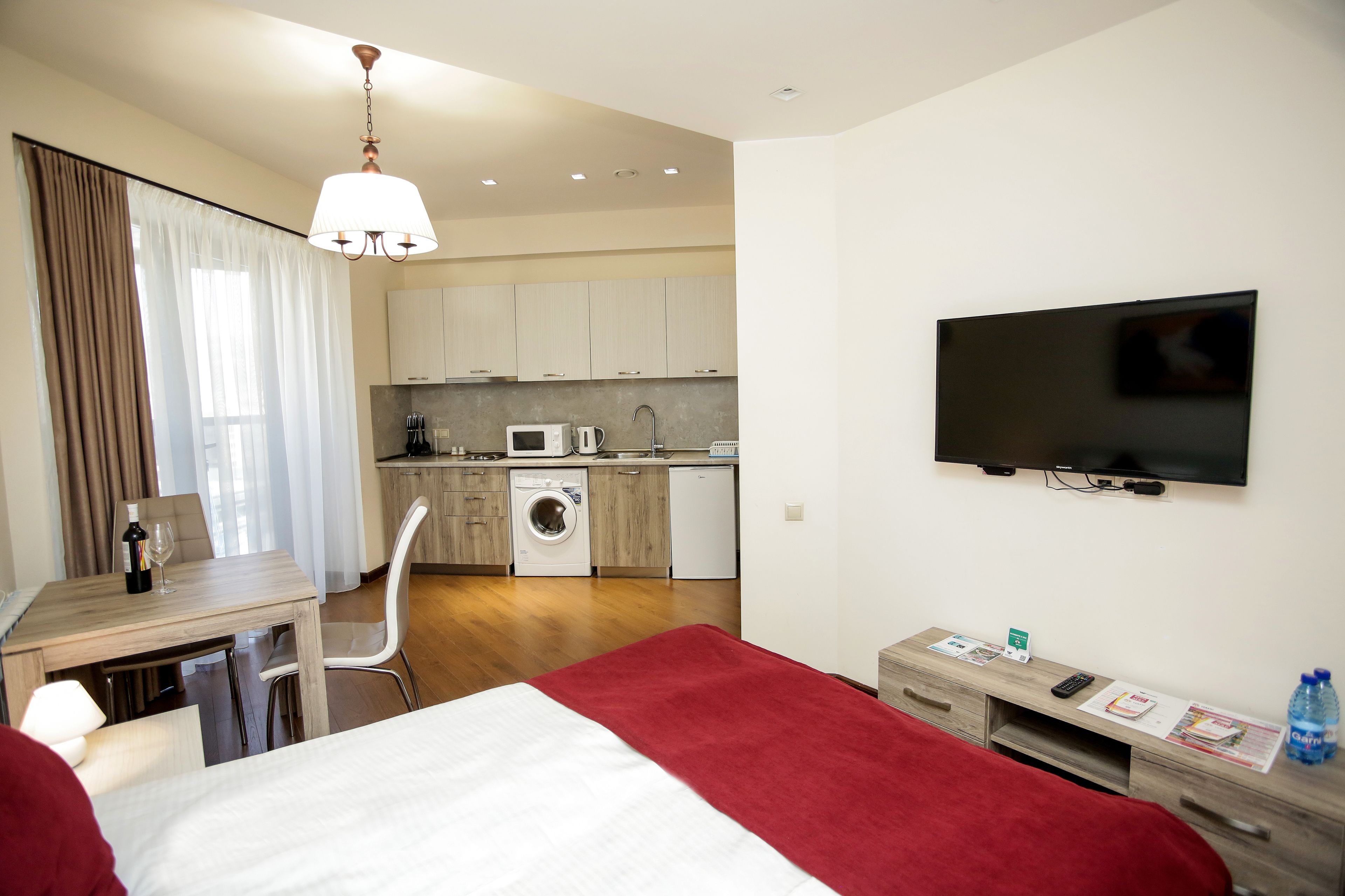 A hotel room with a bed, a desk with a chair, a TV, a small kitchenette, and a washing machine.