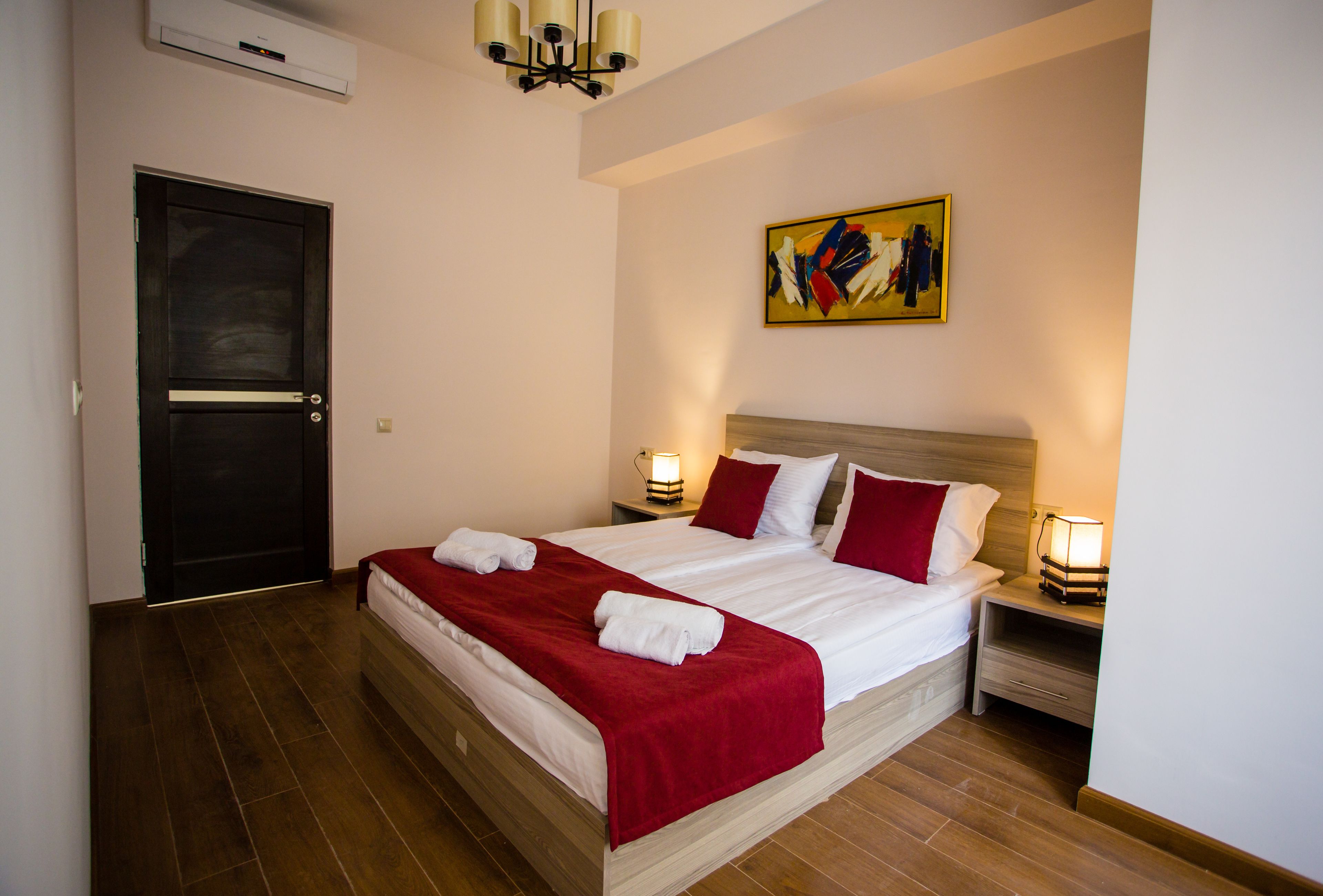 A neatly made bed with white and red linens, two bedside lamps, and a painting on the wall.