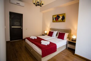 Een net opgemaakt bed met witte en rode linnens, twee bedlampjes en een schilderij aan de muur.