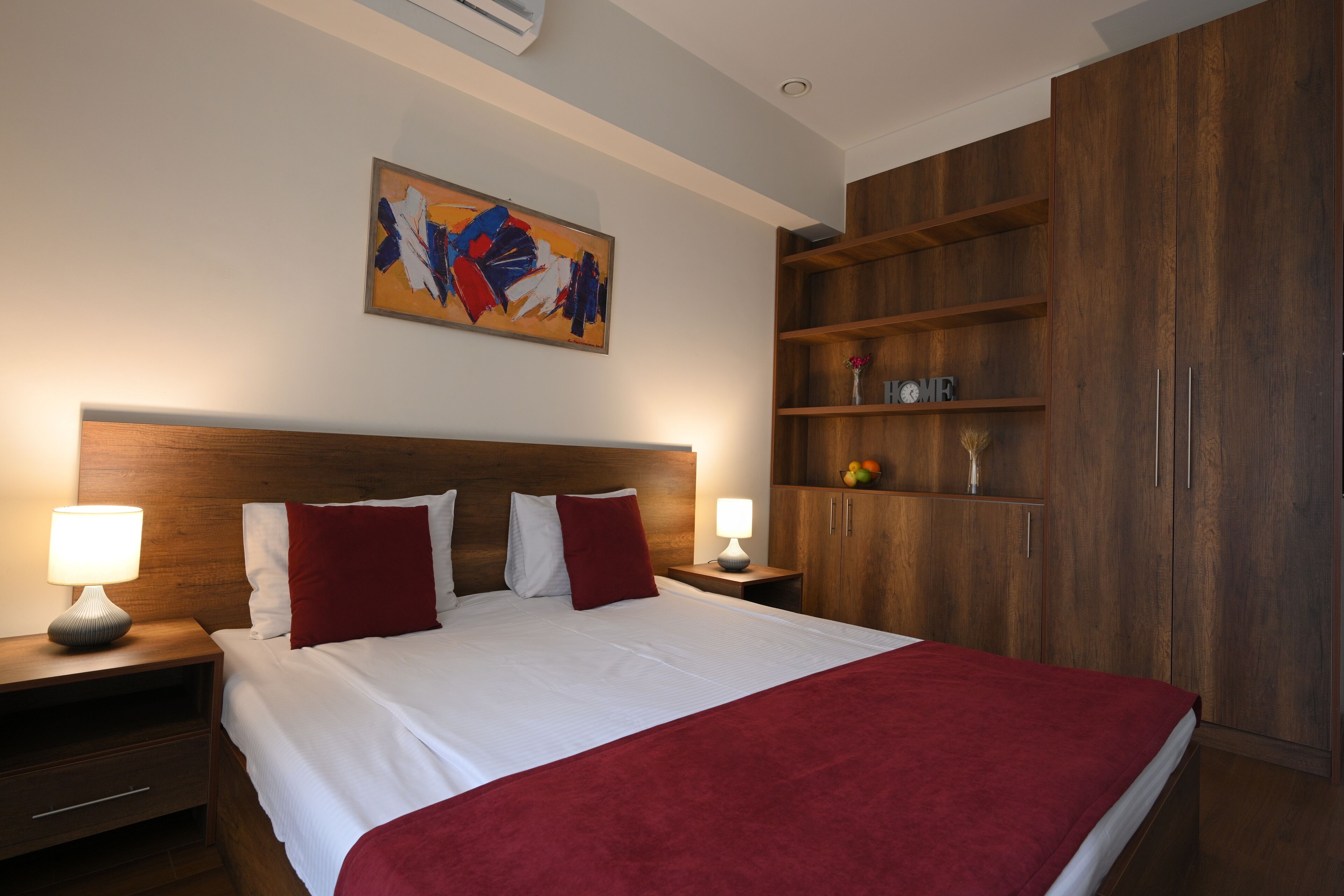 A hotel room with a large bed, wooden headboard, two bedside tables with lamps, a wooden wardrobe, and a colorful abstract painting on the wall.