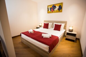 Een keurig opgemaakt bed met rode en witte beddengoed, twee nachtkastjes met lampen, en een kleurrijk abstract schilderij aan de muur.