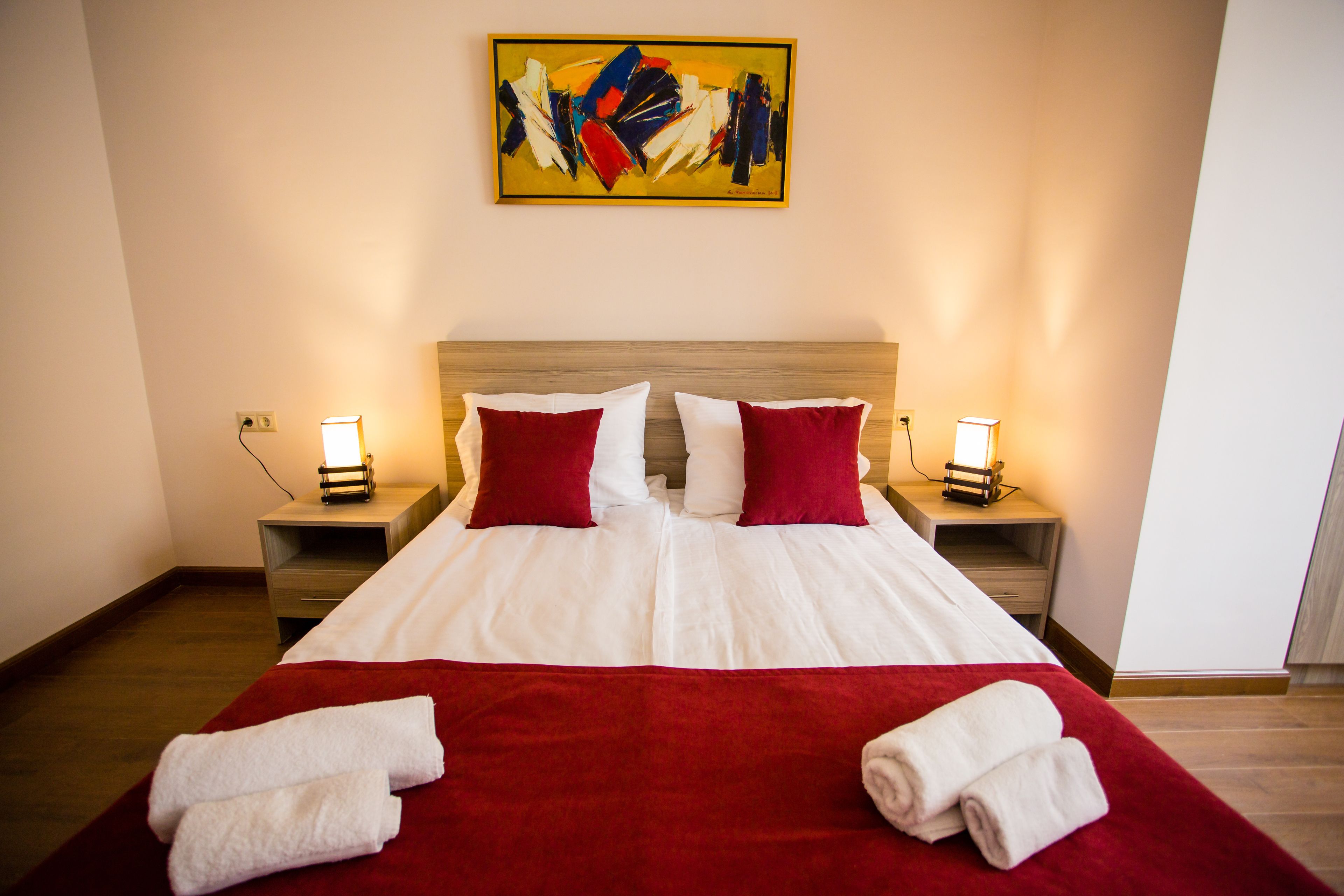 Ein Hotelzimmer mit einem großen Bett, zwei rot-weißen Kissen, einer roten Tagesdecke, zwei Nachttischen mit Lampen und einem Bild an der Wand.