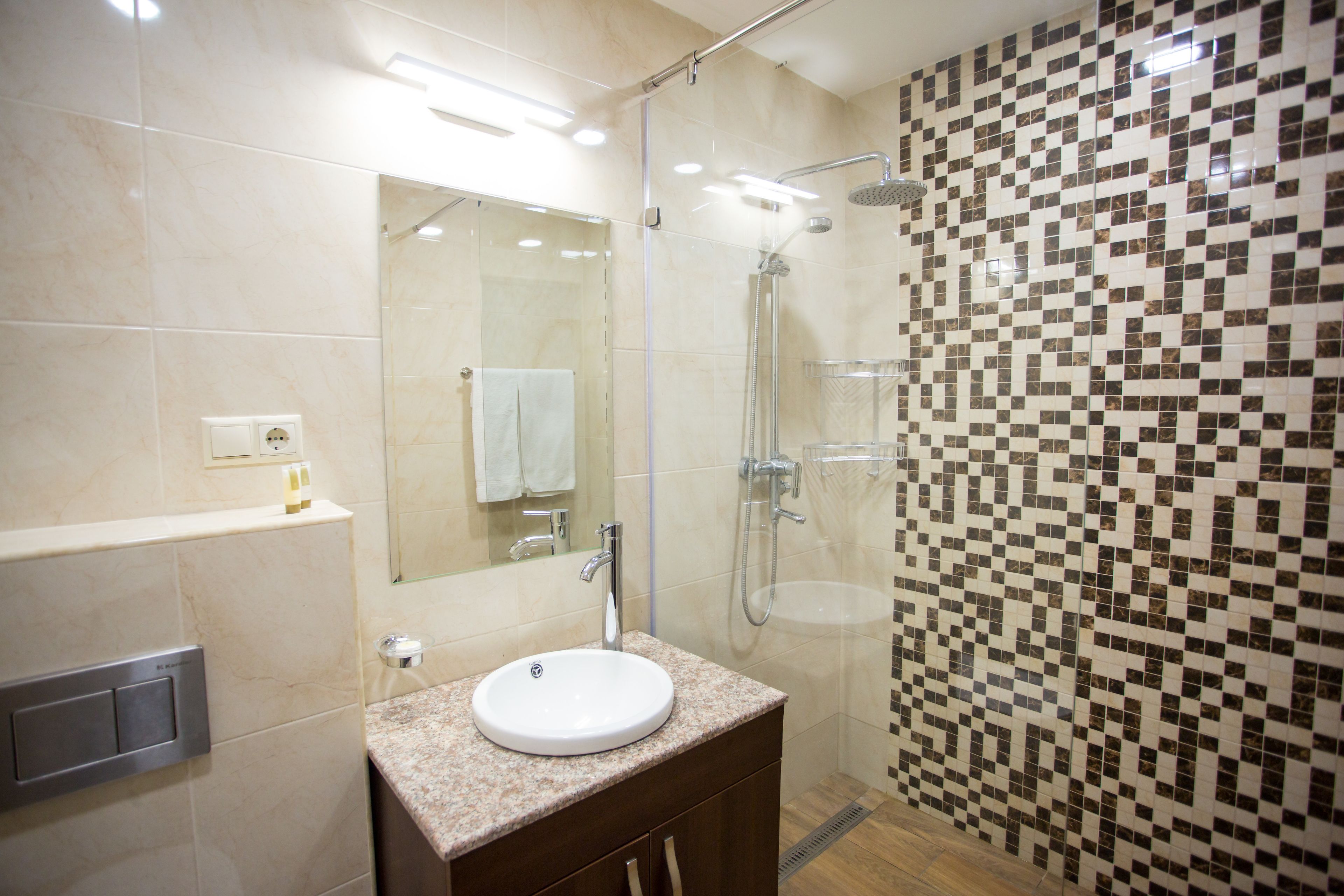 Une salle de bain moderne dotée d’une douche à parois de verre, d’un lavabo avec un plan de travail en marbre et d’un mur recouvert de carreaux formant un motif géométrique.