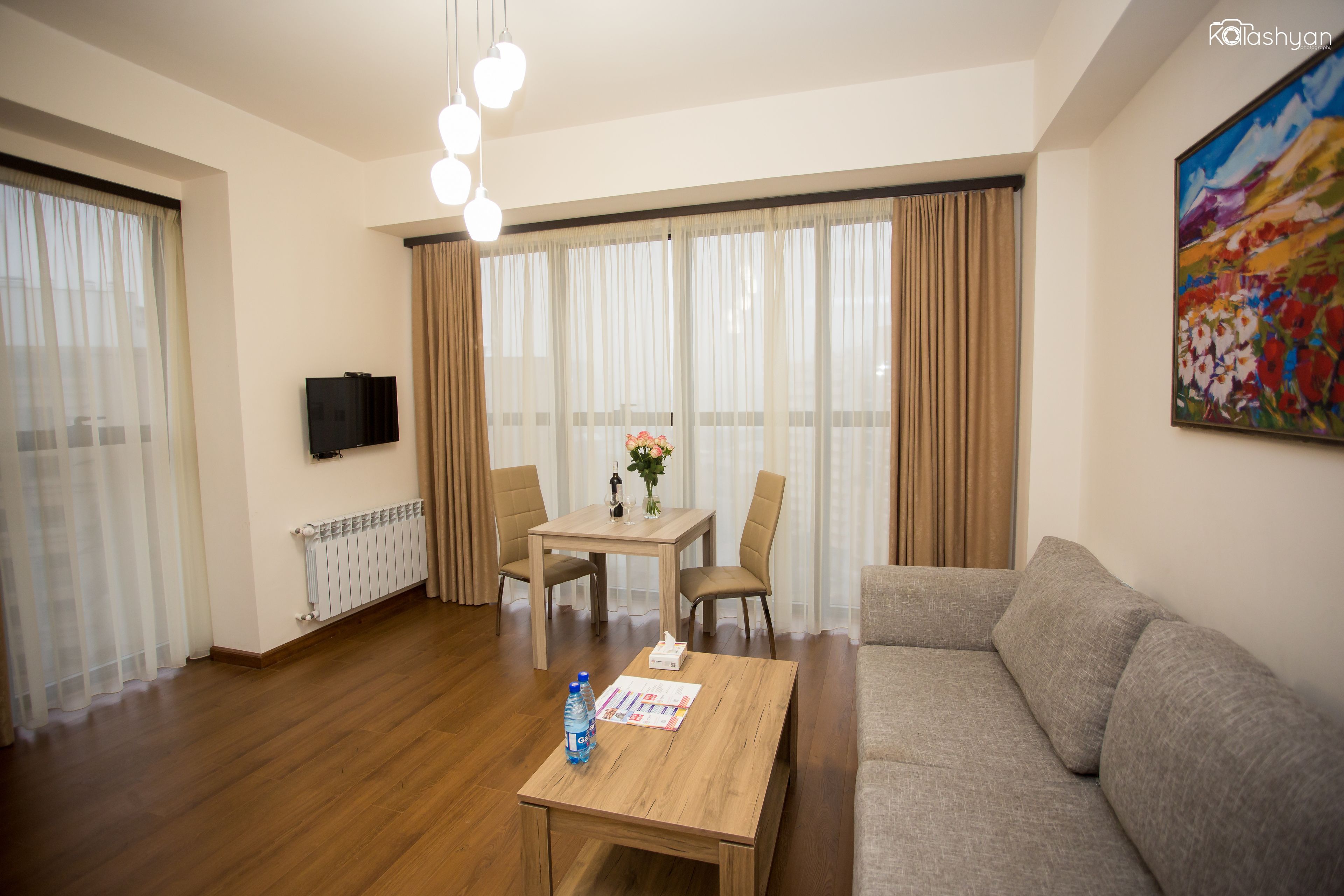 A modern living room with a wooden coffee table, beige sofa, and a dining table with chairs. A flat-screen TV is mounted on the wall, and a painting is visible on the wall.