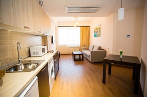 Een knus woongedeelte met een kitchenette, een bank, een eettafel en een groot raam met gordijnen.
