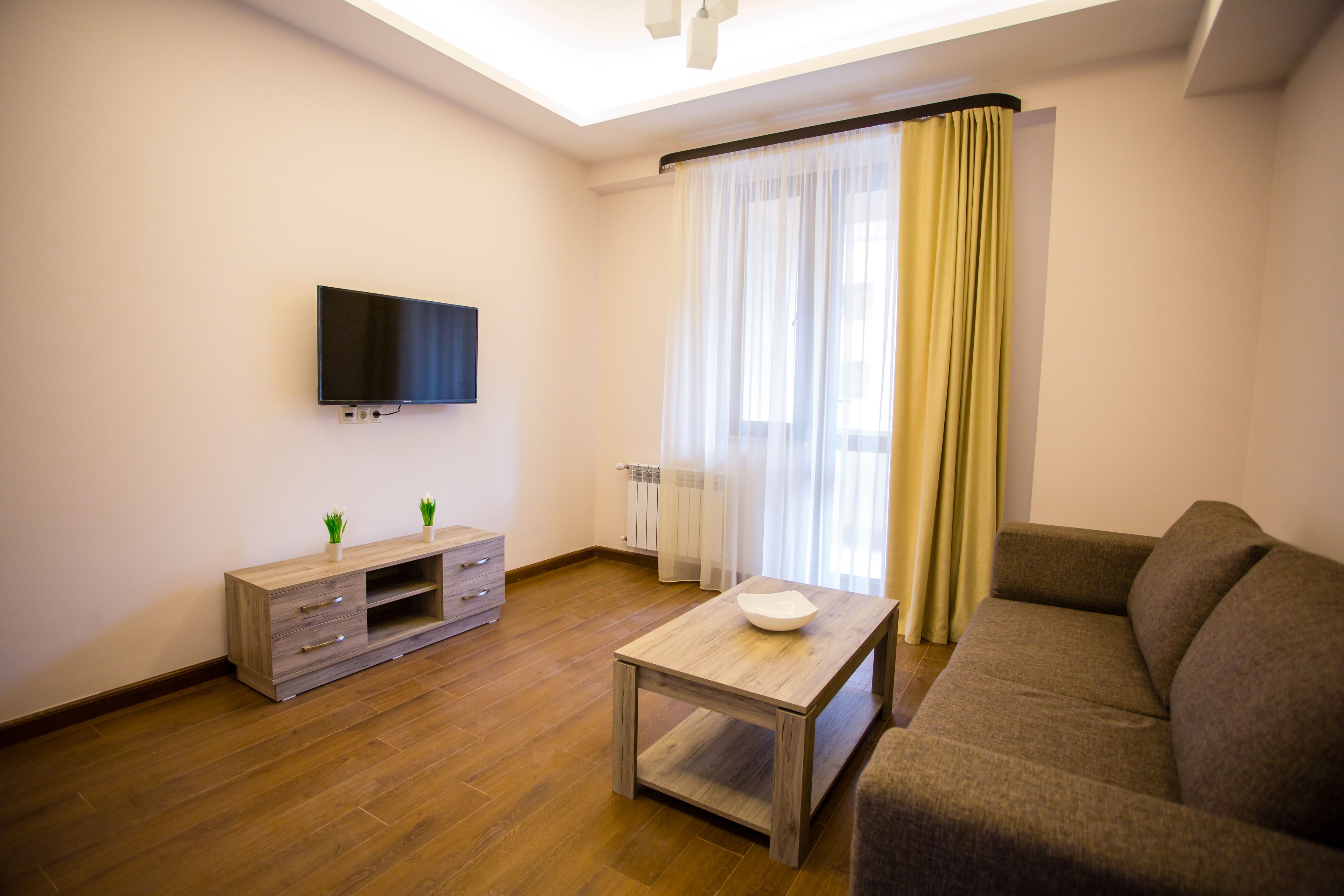 Una sala moderna con un televisor de pantalla plana, una mesa de centro de madera, un sofá, un mueble de televisión con cajones y un ventanal con cortinas translúcidas.