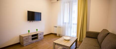 Una sala moderna con un televisor de pantalla plana, una mesa de centro de madera, un sofá, un mueble de televisión con cajones y un ventanal con cortinas translúcidas.