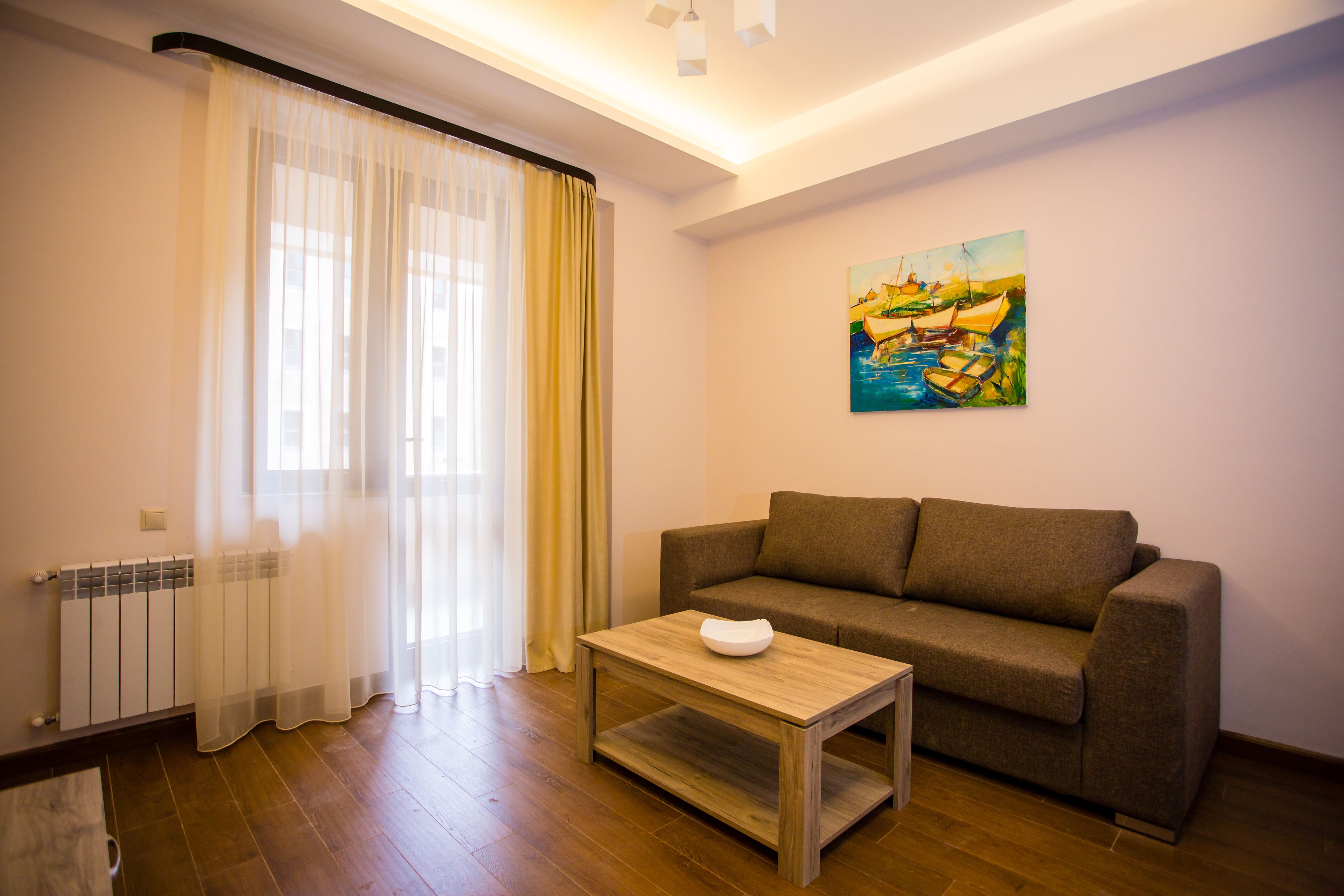 A living room with a brown sofa, a wooden coffee table, a painting on the wall, and a window with sheer curtains.