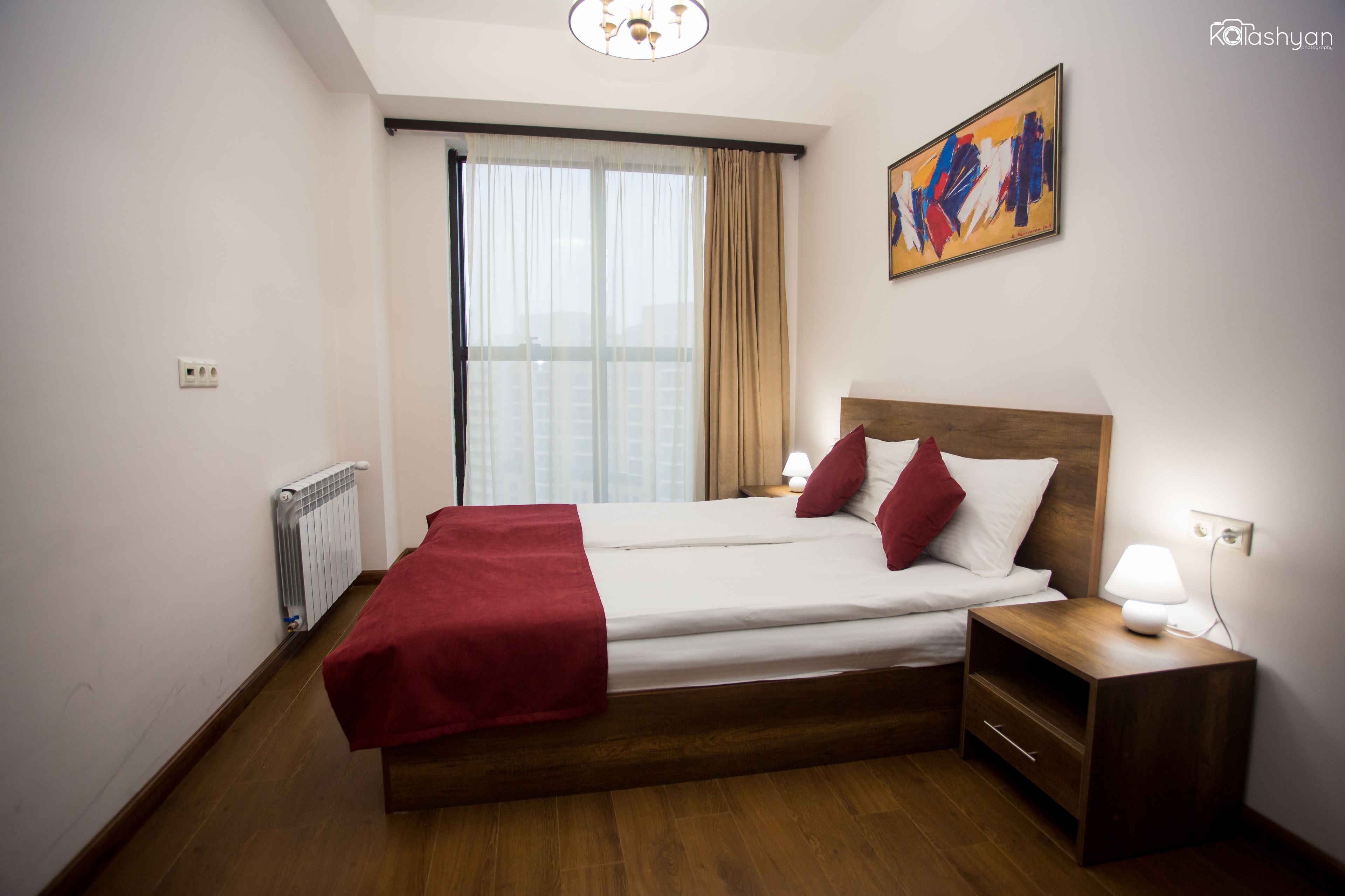 Habitación de hotel con cama, mesita de noche, ventana con cortinas, radiador y un cuadro en la pared.