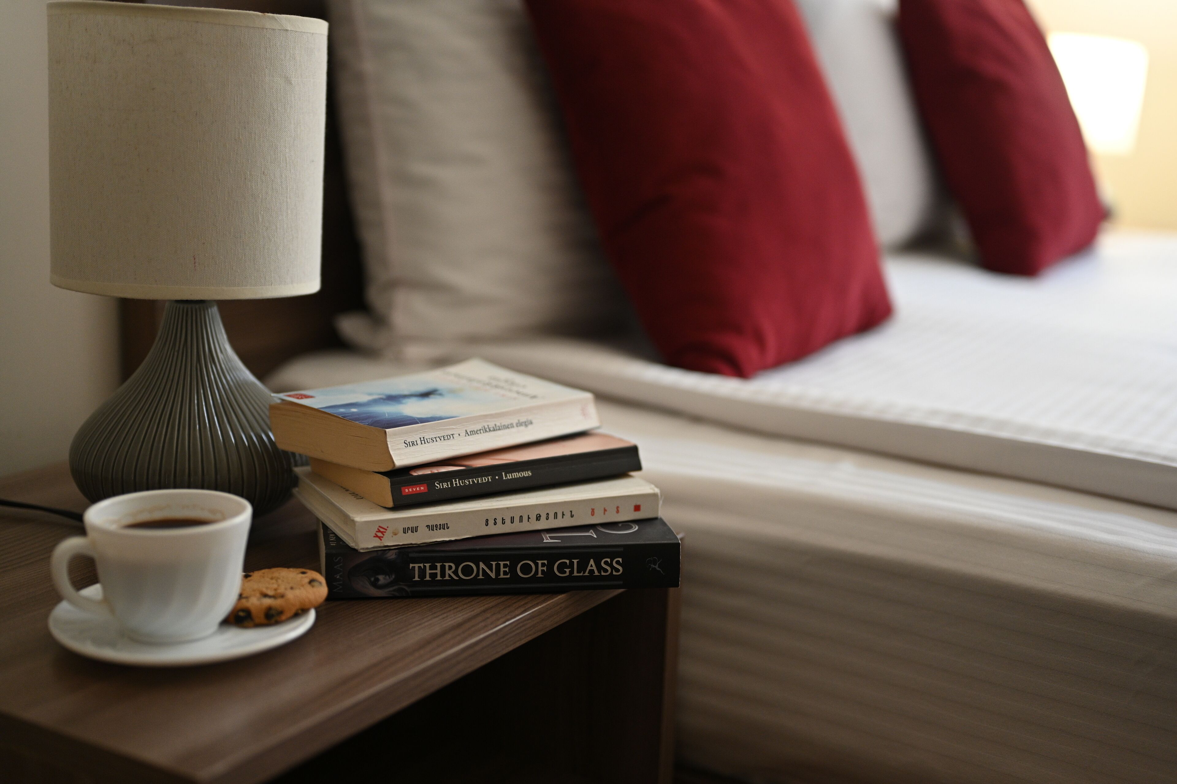 A bedside table with a lamp, a cup of coffee, a cookie, and a stack of books.