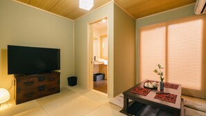 Una sala de estar con un televisor sobre un mueble de madera, una mesa de comedor con individuales y una ventana con persianas.