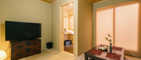 A living room with a TV on a wooden cabinet, a dining table with placemats, and a window with blinds.