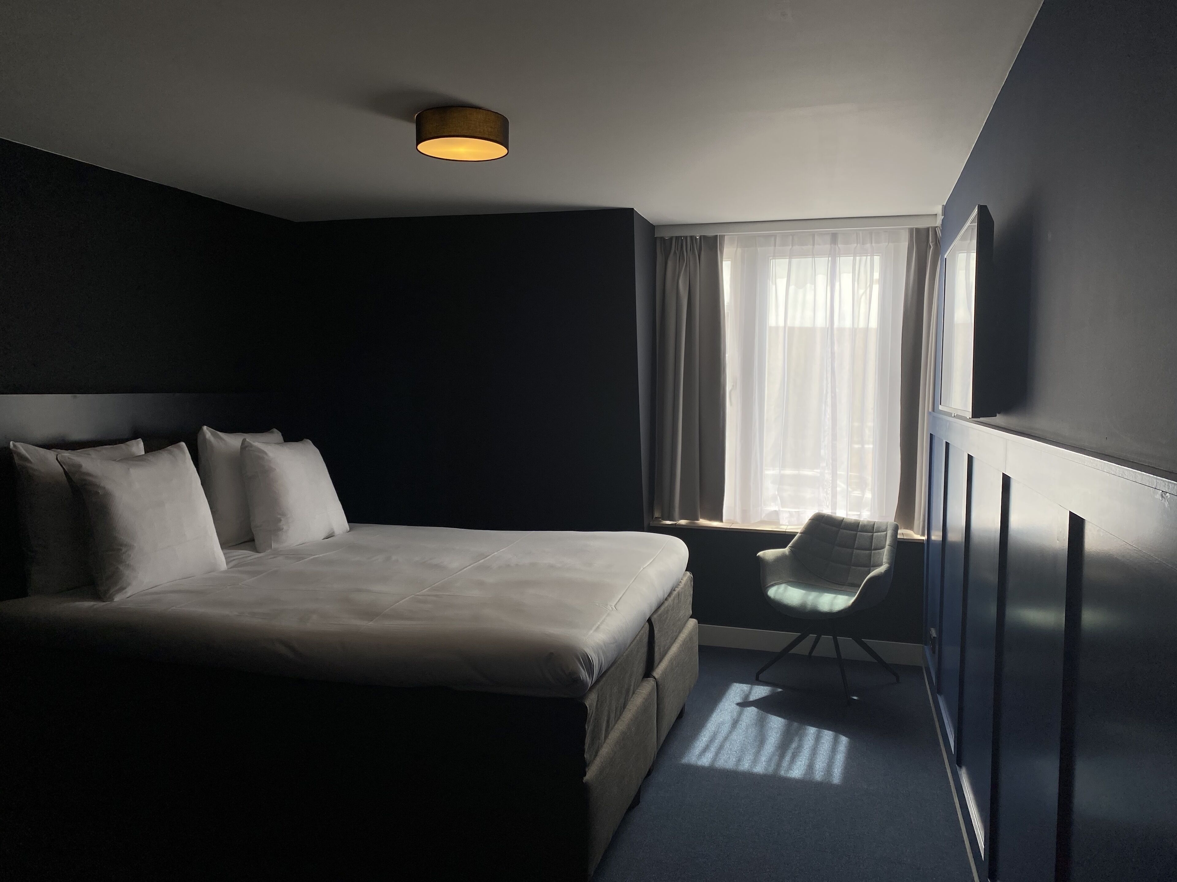 Een moderne hotelkamer met een groot bed, een stoel en een raam met dunne gordijnen.