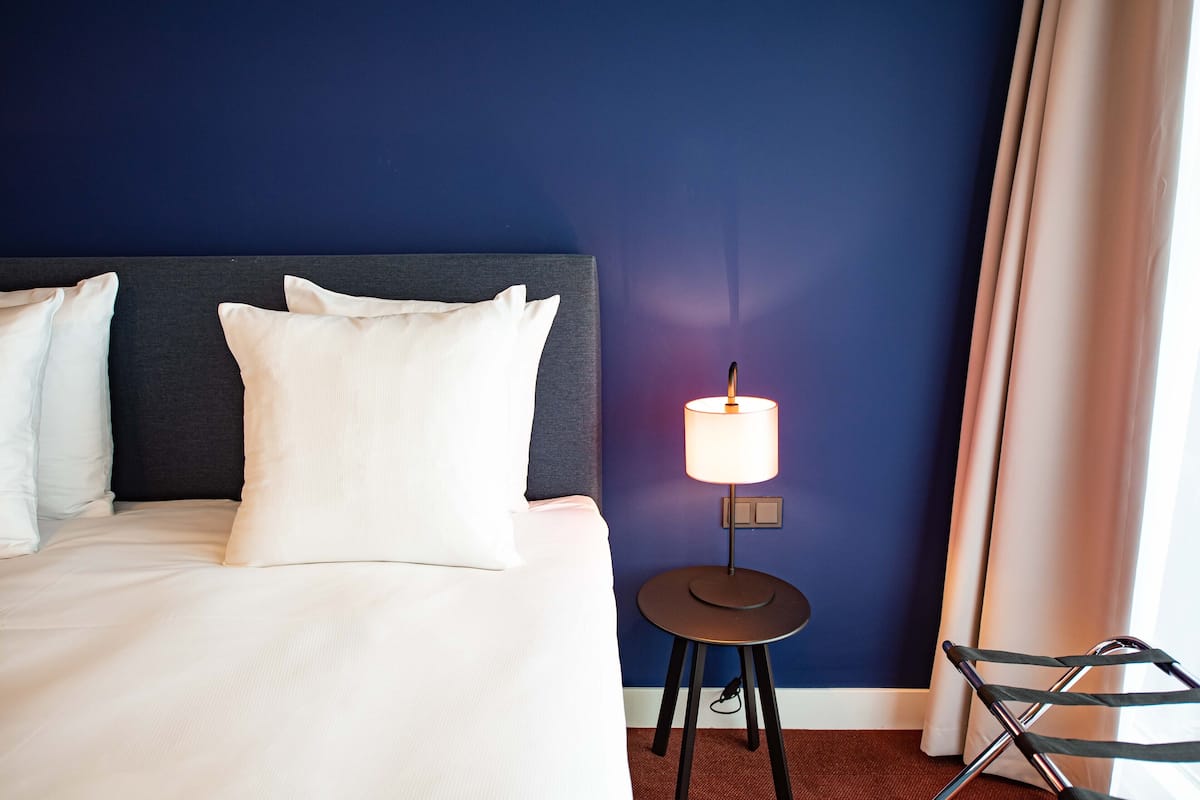 Une chambre d’hôtel avec un lit, une table de chevet avec une lampe, une chaise et une fenêtre avec des rideaux.