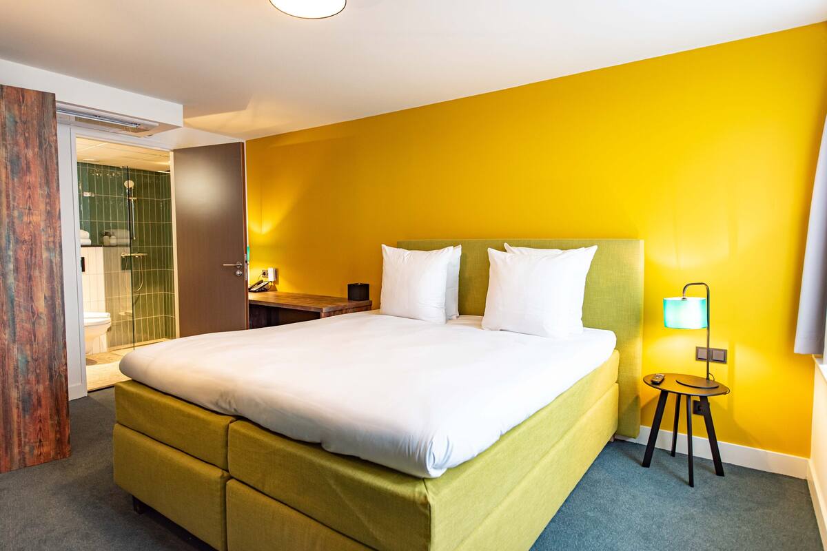 Une chambre d’hôtel avec un grand lit, une table de chevet, une lampe et une salle de bain visible à travers une porte ouverte.