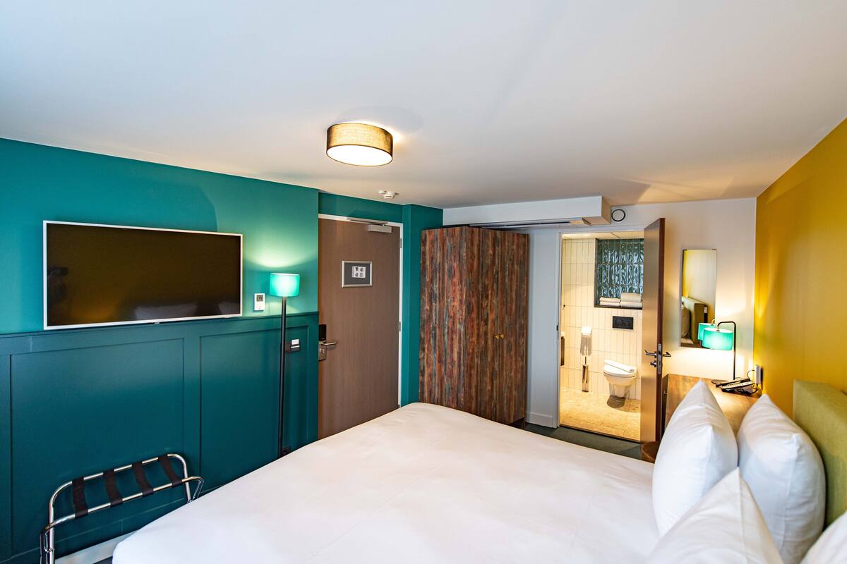 Une chambre d’hôtel aux murs de couleur turquoise, un lit avec du linge de maison blanc, une armoire en bois, une salle de bain avec des toilettes et un lavabo, et un miroir.