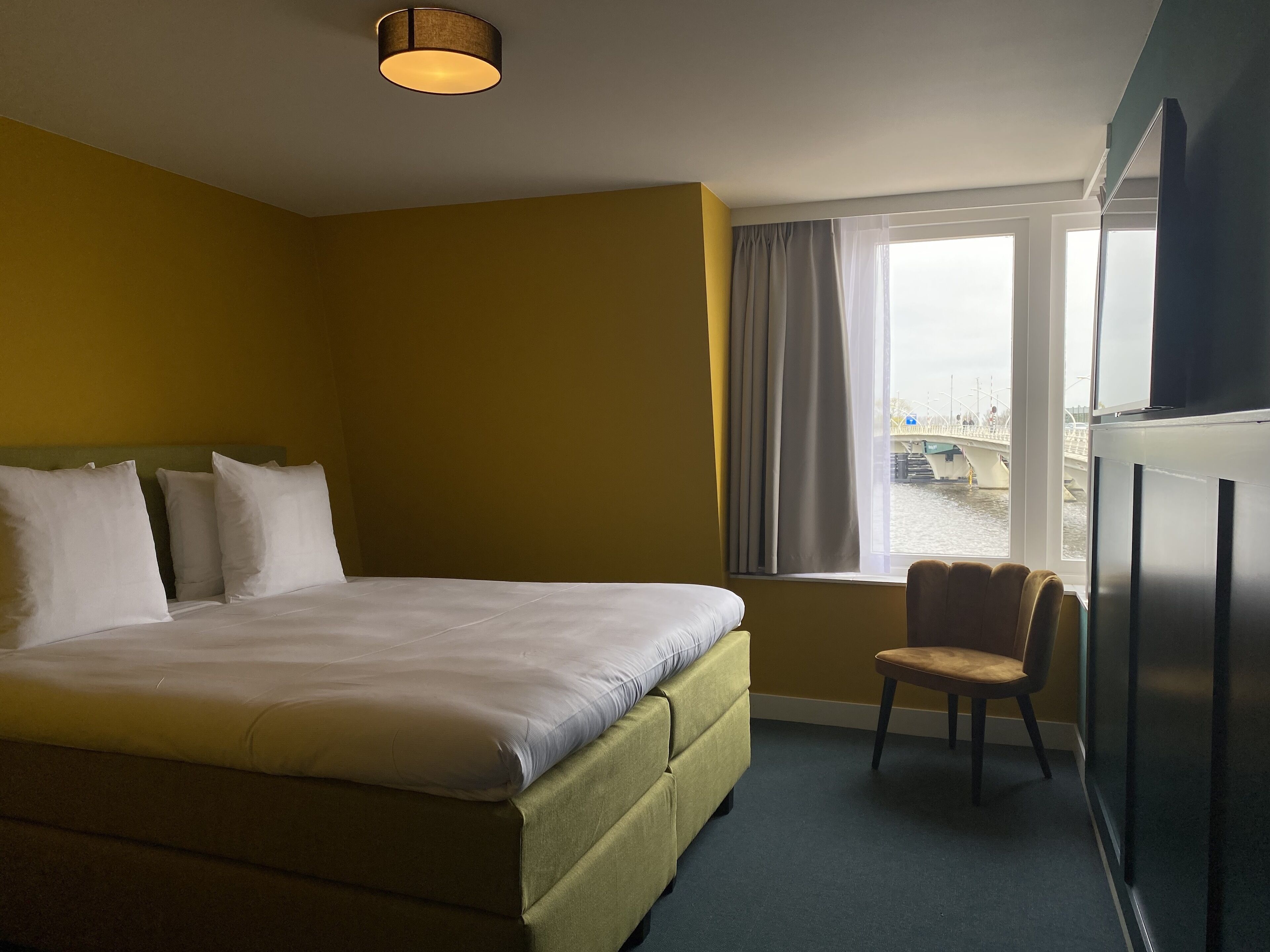 A hotel room with a bed, a chair, a window with curtains, and a view of a bridge and boats outside.