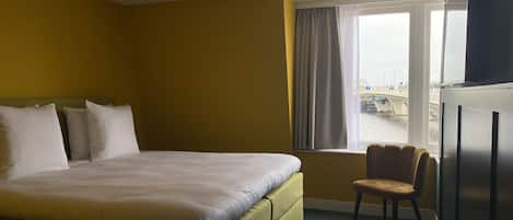 A hotel room with a bed, a chair, a window with curtains, and a view of a bridge and boats outside.