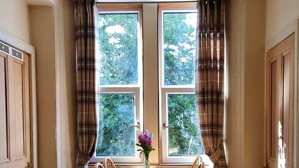 Un coin repas avec une table et des chaises, un vase rempli de fleurs et une vue sur la verdure grâce à de grandes fenêtres.