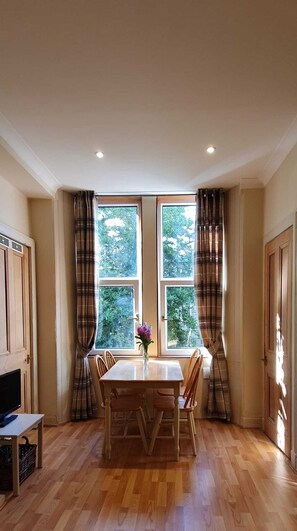 A dining area with a table and chairs, a vase with flowers, and a view of greenery through large windows. - Cosy & Modern Apartment in Central Edinburgh (Edinburgh)