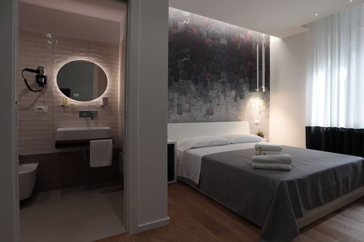 A modern bedroom with a large bed, a bedside table, a mirror, and a window with curtains.