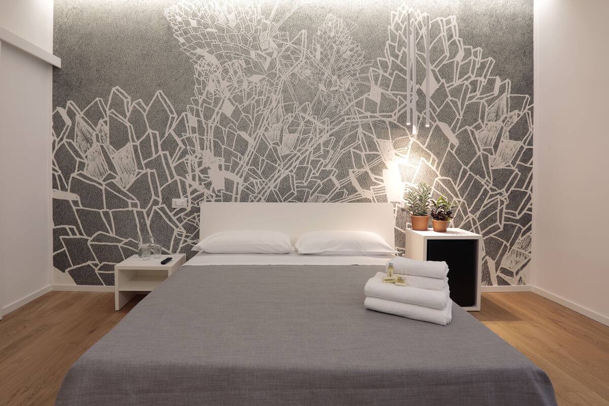 A modern bedroom with a grey bedspread, white pillows, a bedside table with a remote control, a nightstand, and a wall with a geometric pattern.