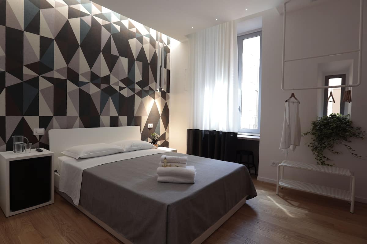 A modern bedroom with a geometric-patterned wall, a large bed, bedside tables, a hanging plant, and a view of the outside through a window.