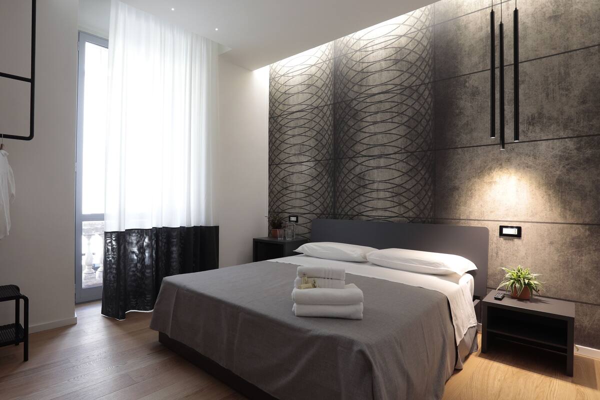 A modern bedroom with a large bed, a bedside table with a lamp, a small potted plant, and a view of the outdoors through a window with sheer curtains.