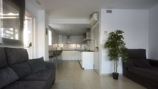A modern living room with a sofa, a dining area with chairs, and a kitchen with a central island. - Residencia Universitaria Alboran (Málaga)