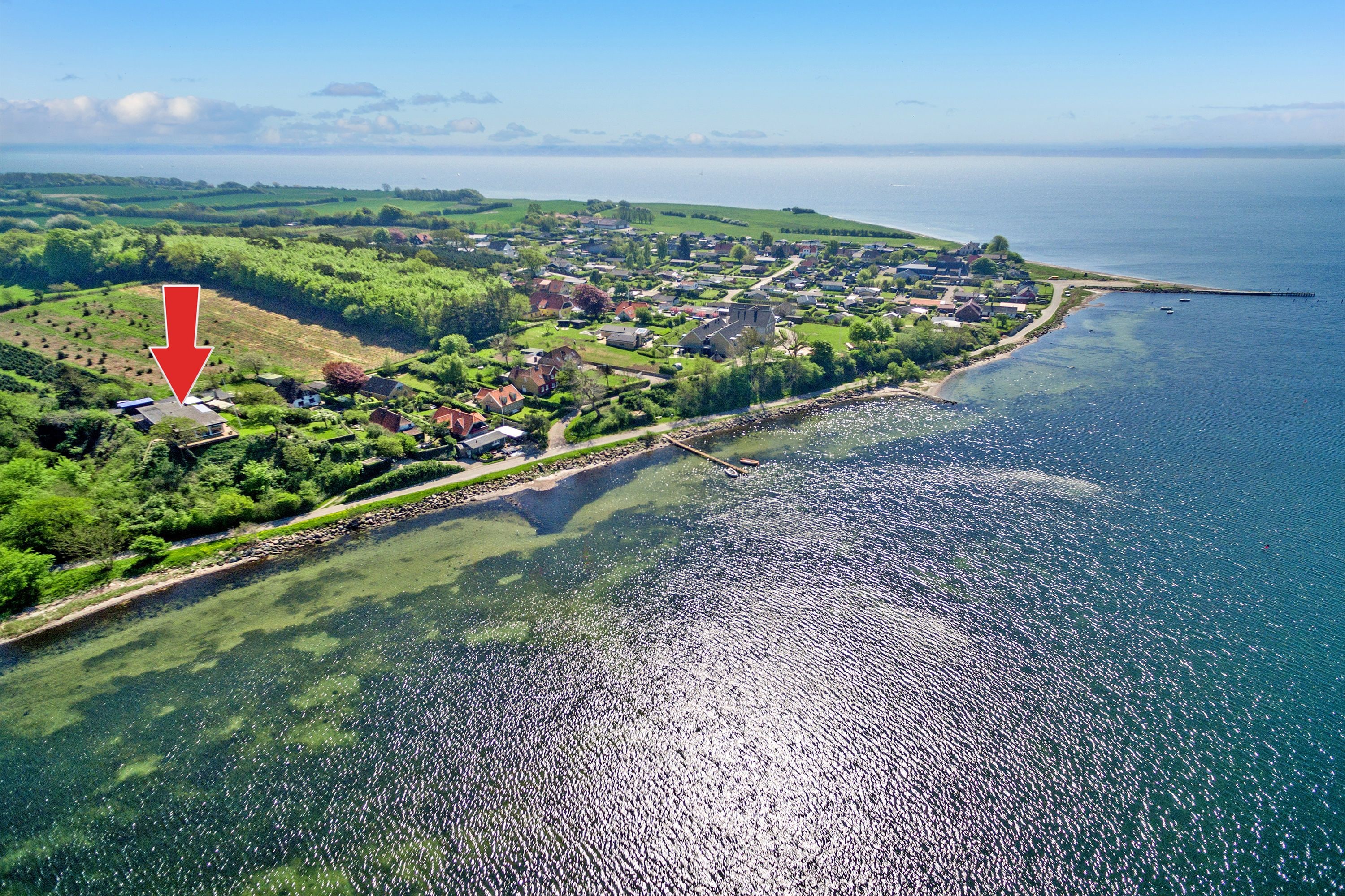 Une vue aérienne d’une ville côtière, avec une vue dégagée sur la mer, des collines verdoyantes et une flèche rouge indiquant un endroit précis.