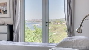 A bedroom with a large window, a bed, a bedside table, and a lamp.