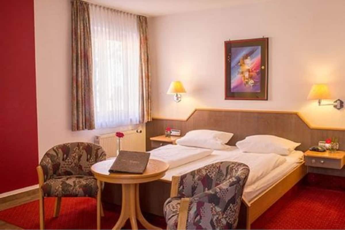 Une chambre d’hôtel avec un lit, deux chaises, une petite table, une lampe, un tableau et une fenêtre avec des rideaux.