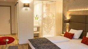 Ein modernes Hotelzimmer mit einem großen Bett, einem Nachttisch und einem Beistelltisch. Ein Badezimmer ist durch eine Glastür sichtbar.